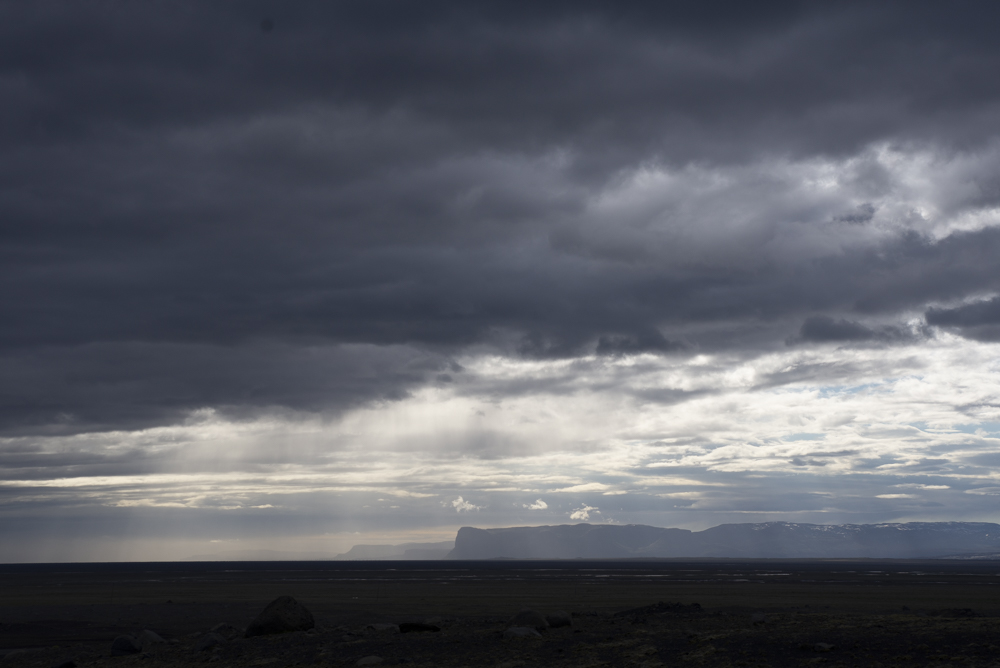 Distant escarpment across a black volcanic plain with sunbeams mixing with rain under a dramatic sky, captured near Eldhraun, Iceland during our June 2016 trip.